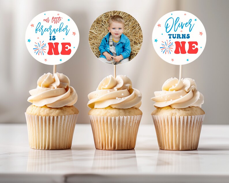 May include: Three cupcakes with white frosting and red and blue sprinkles topped with round paper cupcake toppers. The toppers feature a photo of a young boy, the text "our little firecracker is one" and the text "Oliver turns one".