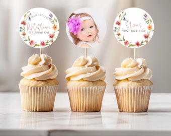 May include: Three cupcakes with white frosting and sprinkles topped with round floral themed paper cupcake toppers. One topper has a photo of a baby girl with a pink flower headband, another says "Our Little Wildflower is turning one", and the third says "Olivia's First Birthday".