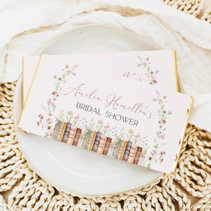 Puede incluir: Una invitación rosa claro con bordes dorados, con el texto "Amelia Hamilton's Bridal Shower". El diseño incluye ilustraciones de acuarela de flores y una fila de libros. La invitación está colocada sobre un plato blanco.