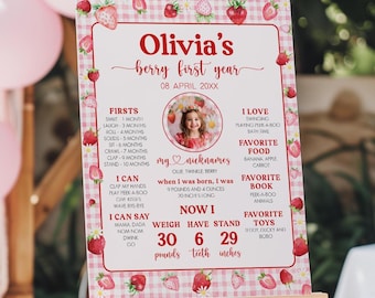 May include: A wooden easel displays a sign with a strawberry theme. The sign reads "Olivia's berry first year" with details about the child's milestones, weight, and height. The sign is decorated with strawberries and a gingham pattern.