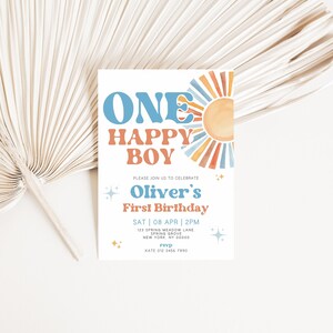 以下が含まれることがあります： 青とオレンジ色の「ONE HAPPY BOY」の文字と水彩画の太陽のイラストが描かれた白い招待状。カードには「Oliver's First Birthday」と日付と時間の詳細が記載されています。背景には、乾燥したヤシの葉の扇子があります。
