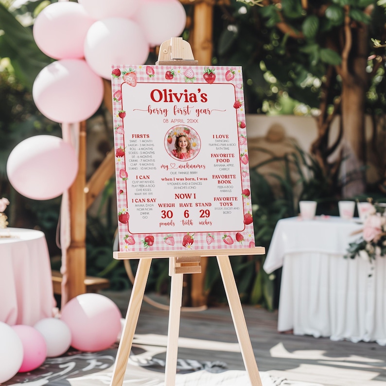 May include: A pink and white gingham and strawberry-themed first birthday sign with a photo of a girl. The sign includes the girl's name, Olivia, and her birthdate. It also lists her firsts, favorites, and current stats, including her weight, height, and number of teeth.