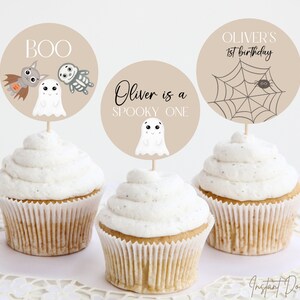 Puede incluir: Tres cupcakes con adornos beige con dise&ntilde;os de Halloween. Uno dice "BOO" con un murci&eacute;lago, un fantasma y un esqueleto. Otro dice "Oliver is a spooky one" con un fantasma. El tercero dice "Oliver's 1st birthday" con una ara&ntilde;a y una telara&ntilde;a.