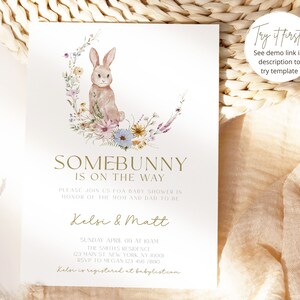 May include: A baby shower invitation featuring a watercolour illustration of a bunny rabbit sitting on a crescent moon surrounded by flowers. The text reads "Somebunny is on the way" and includes the names Kelsi & Matt, the date and time of the shower, and the address.