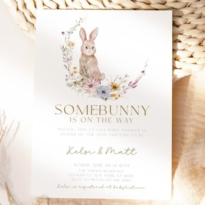 May include: A baby shower invitation featuring a watercolour illustration of a bunny rabbit sitting in a wreath of flowers. The text reads "Somebunny is on the way" and includes the names "Kelsi & Matt".
