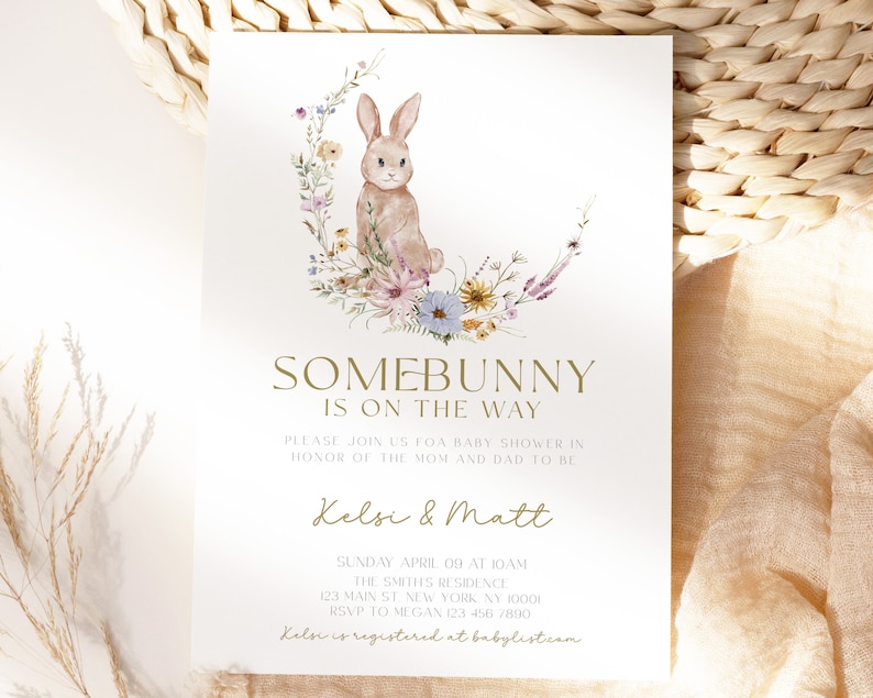 May include: A baby shower invitation featuring a watercolour illustration of a bunny rabbit sitting in a wreath of flowers. The text reads "Somebunny is on the way" and includes the names "Kelsi & Matt".