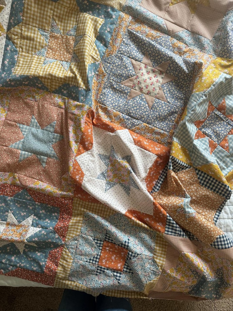 May include: A patchwork quilt with a variety of floral and geometric patterns in shades of blue, orange, and yellow. The quilt is made up of squares and stars, and it is folded in half.