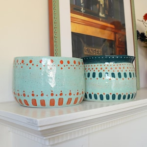 May include: Two ceramic bowls with a speckled mint green glaze. The left bowl features orange dots and vertical dashes. The right bowl has a dark teal rim, orange dots, and dark teal vertical dashes. Both bowls have a smooth, glossy finish.