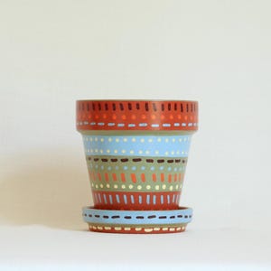 May include: A terracotta plant pot with a matching saucer, featuring horizontal bands of hand-painted patterns in light blue, olive green, orange, and dark brown. The design consists of repeating dots and dashes encircling the exterior surface.