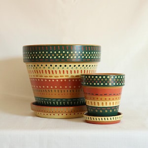 May include: Two hand-painted terracotta flower pots with matching saucers. The larger pot is 8 inches in diameter and 7 inches tall, while the smaller pot is 4 inches in diameter and 4 inches tall. Both pots feature a colorful design with green, red, yellow, and brown bands.