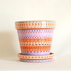 May include: A hand-painted terracotta plant pot with a matching saucer, featuring horizontal bands of light purple, peach, and orange. The surface is decorated with repeating patterns of small dots and dashes in blue, dark purple, and orange paint.