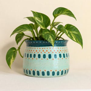 May include: A ceramic planter with a light mint green base featuring dark teal vertical dashes and small orange dots. The rim is a solid dark teal with a row of white dots. A pothos plant with variegated green and yellow heart-shaped leaves grows inside.