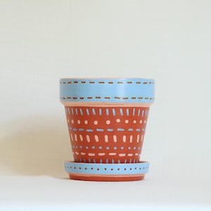 May include: A terracotta plant pot with a matching saucer, featuring a light blue painted rim and base with a pattern of dots and dashes in light blue and white across the reddish-brown surface of the pot and the light blue saucer.
