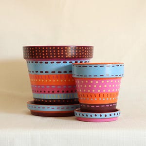 May include: Two hand-painted terracotta flower pots with matching saucers. The larger pot is blue, orange, and pink with a brown rim. The smaller pot is pink, orange, and blue with a brown rim. Both pots have dotted and dashed patterns.