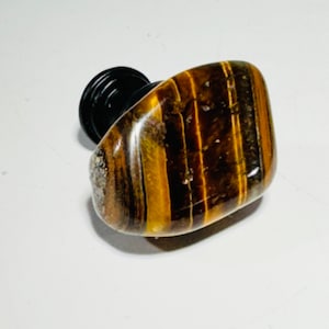 May include: A polished tiger's eye stone attached to a black base. The stone is a mix of brown and gold with vertical stripes. The base is round and made of black plastic. The stone is smooth and has a glossy finish.