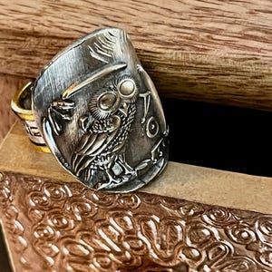 May include: A silver ring with an owl design. The owl is wearing a top hat and has large, round eyes. The ring is set on a wooden surface.