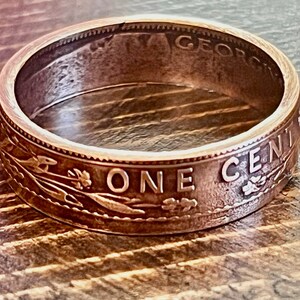 May include: A copper-colored ring crafted from a vintage one-cent coin. The ring features the words "ONE CENT" and "GEORGIVS" along the edges, with detailed engravings of wheat stalks and other designs. The ring is set against a wooden surface.