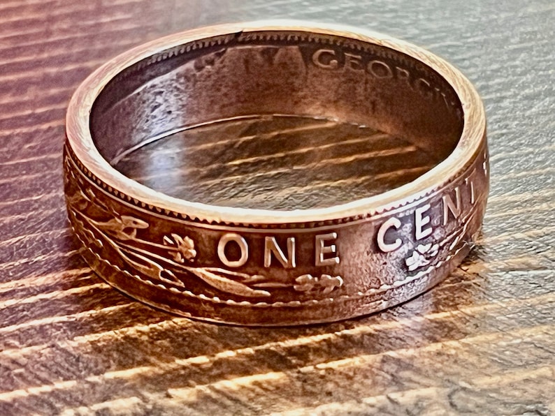 May include: A copper-colored ring crafted from a vintage one-cent coin. The ring features the words "ONE CENT" and "GEORGIVS" along the edges, with detailed engravings of wheat stalks and other designs. The ring is set against a wooden surface.