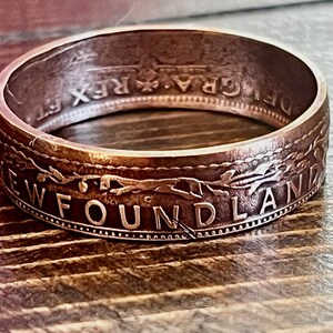 May include: A close-up of a ring made from a coin. The ring is a copper color with the word "NEWFOUNDLAND" engraved on the side. The ring has a detailed design and is set against a wooden surface.