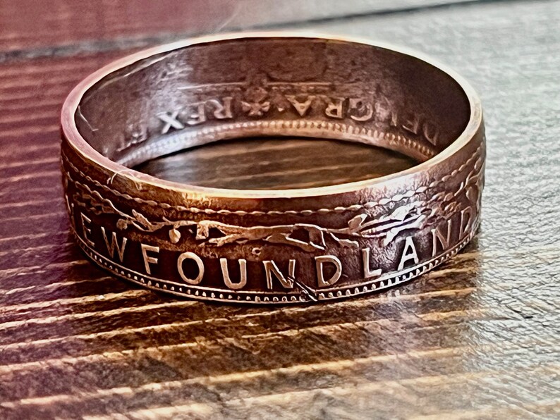 May include: A close-up of a ring made from a coin. The ring is a copper color with the word "NEWFOUNDLAND" engraved on the side. The ring has a detailed design and is set against a wooden surface.