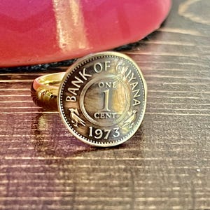 May include: A gold-colored ring featuring a vintage 1973 one-cent coin from the Bank of Guyana. The coin has text that reads "BANK OF GUYANA ONE 1 CENT 1973" and is set in a simple gold band.