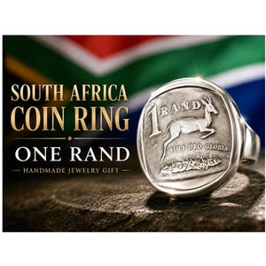 May include: A silver coin ring featuring a South African one rand coin. The coin shows a leaping springbok and the text "1 RAND" and "SOLI DEO GLORIA". The ring is set against the South African flag.