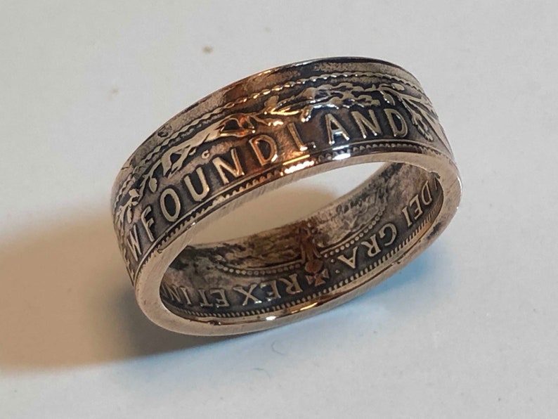 May include: A silver ring with the word "NEWFOUNDLAND" engraved on the outer band. The ring has intricate detailing and text, with a vintage or antique appearance. The ring is a unique piece of jewelry.