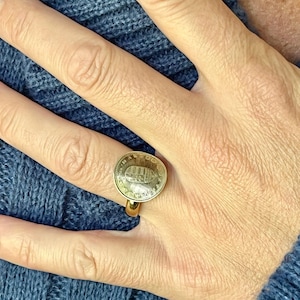 May include: A gold-colored ring featuring a coin with an embossed design. The ring is worn on a hand, with a blue knitted sweater in the background. The coin has a detailed image, and the ring band is smooth.