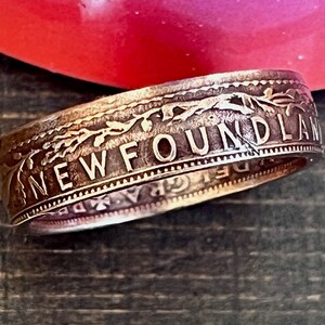 May include: A silver ring with the word "NEWFOUNDLAND" engraved on the outer band. The ring has a detailed floral design and appears to be made from a coin. The ring is set against a blurred background.