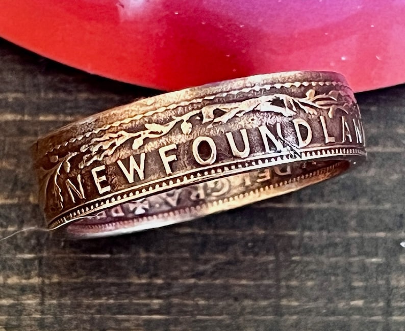 May include: A silver ring with the word "NEWFOUNDLAND" engraved on the outer band. The ring has a detailed floral design and appears to be made from a coin. The ring is set against a blurred background.