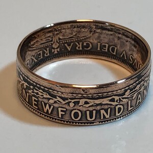 May include: A copper-colored ring with the word "NEWFOUNDLAND" engraved around the band. The ring has a detailed design with text and patterns. The ring is likely made from a coin, with visible text and imagery.