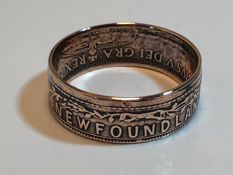 May include: A copper-colored ring with the word "NEWFOUNDLAND" engraved around the band. The ring has a detailed design with text and patterns. The ring is likely made from a coin, with visible text and imagery.