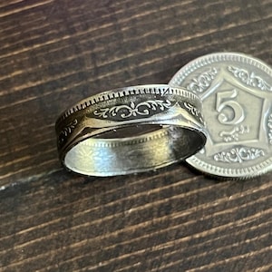 Puede incluir: Un anillo de plata con un diseño floral ornamentado y una moneda con el número 5. El anillo tiene un patrón detallado alrededor de su banda. La moneda está al lado del anillo.