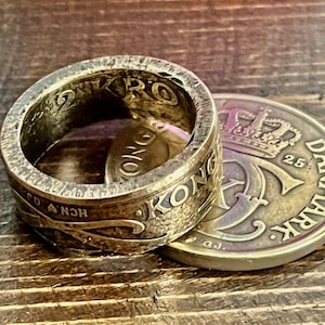 Könnte beinhalten: Ein silberner Ring aus einer Münze, mit dem Wort "KONGE" sichtbar. Daneben liegt eine dänische Münze mit einer Krone und dem Wort "DANMARK". Der Ring hat ein abgenutztes, strukturiertes Aussehen.
