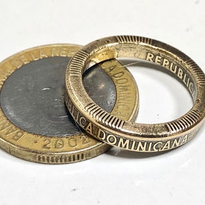 Può includere: Un anello color oro con le parole "REPUBLICA DOMINICANA" incise sulla fascia esterna, accanto a una moneta della Repubblica Dominicana. L'anello ha una serie di linee verticali sui bordi. La moneta è datata 2002.