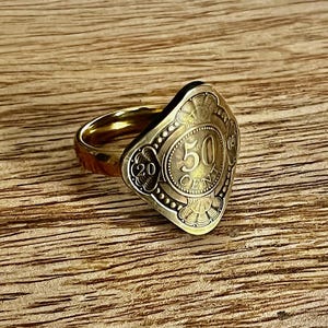 Może przedstawiać: Złoty pierścionek z dużym, diamentowym wzorem monety. Moneta zawiera liczbę "50" i słowo "CENT" w wypukłych literach.