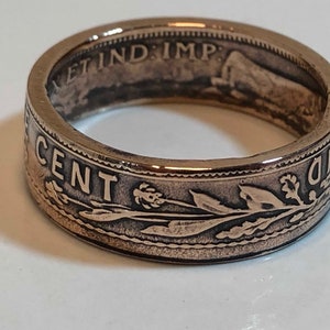 May include: A bronze-colored ring with etched details. The ring features the words "ONE CENT" and other inscriptions. The design includes floral and leaf patterns. The ring is a close-up shot, showcasing the intricate details.
