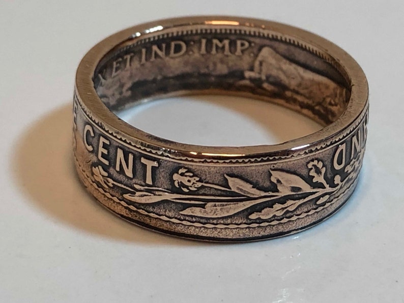 May include: A bronze-colored ring with etched details. The ring features the words "ONE CENT" and other inscriptions. The design includes floral and leaf patterns. The ring is a close-up shot, showcasing the intricate details.