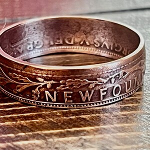 May include: A silver-toned ring with engraved text and floral designs. The ring's outer band features the word "NEWFOUND" and other inscriptions. The ring is set against a wooden surface, highlighting its metallic sheen.