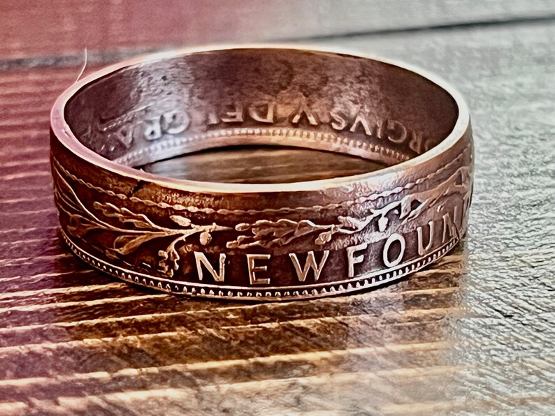 May include: A silver-toned ring with engraved text and floral designs. The ring's outer band features the word "NEWFOUND" and other inscriptions. The ring is set against a wooden surface, highlighting its metallic sheen.