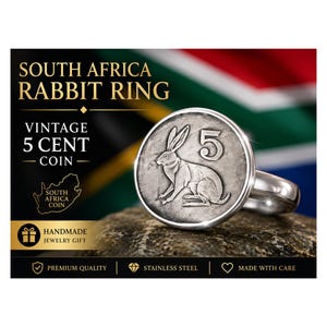 May include: A silver-toned ring featuring a vintage South African 5-cent coin with a rabbit design. The ring is set against a backdrop of the South African flag and includes text that reads "South Africa Rabbit Ring".
