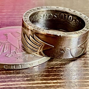 Op de afbeelding: Een zilverkleurige ring met een adelaarontwerp en de inscriptie "8. DEUTSCHEMARK". De ring ligt naast een munt met een adelaarembleem en het jaar 1973.