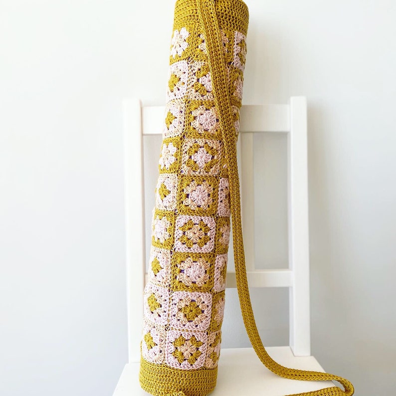 K&ouml;nnte beinhalten: Ein gelber und rosa geh&auml;kelter Yogamatte-Tr&auml;ger mit einem langen Riemen. Der Tr&auml;ger besteht aus Granny Squares und hat ein schlichtes, modernes Design.