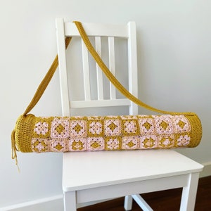 K&ouml;nnte beinhalten: Eine geh&auml;kelte Yogamatte-Tasche in Senfgelb und Rosa. Die Tasche hat einen langen Riemen und steht auf einem wei&szlig;en Stuhl.