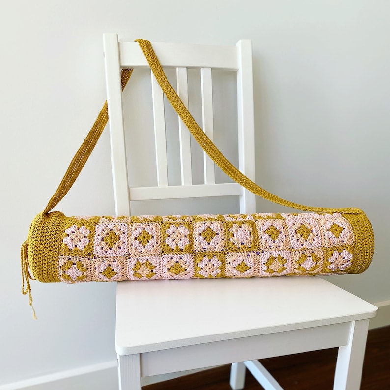 K&ouml;nnte beinhalten: Eine geh&auml;kelte Yogamatte-Tasche in Senfgelb und Rosa. Die Tasche hat einen langen Riemen und steht auf einem wei&szlig;en Stuhl.
