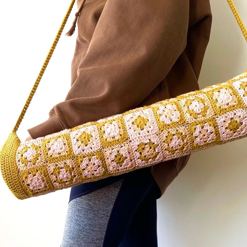 K&ouml;nnte beinhalten: Eine geh&auml;kelte Yogamatte-Tasche mit einem langen Riemen, die ein geometrisches Muster in Rosa und Senfgelb aufweist. Die Tasche wird &uuml;ber der Schulter einer Person getragen, die einen braunen Pullover und eine graue Hose tr&auml;gt.