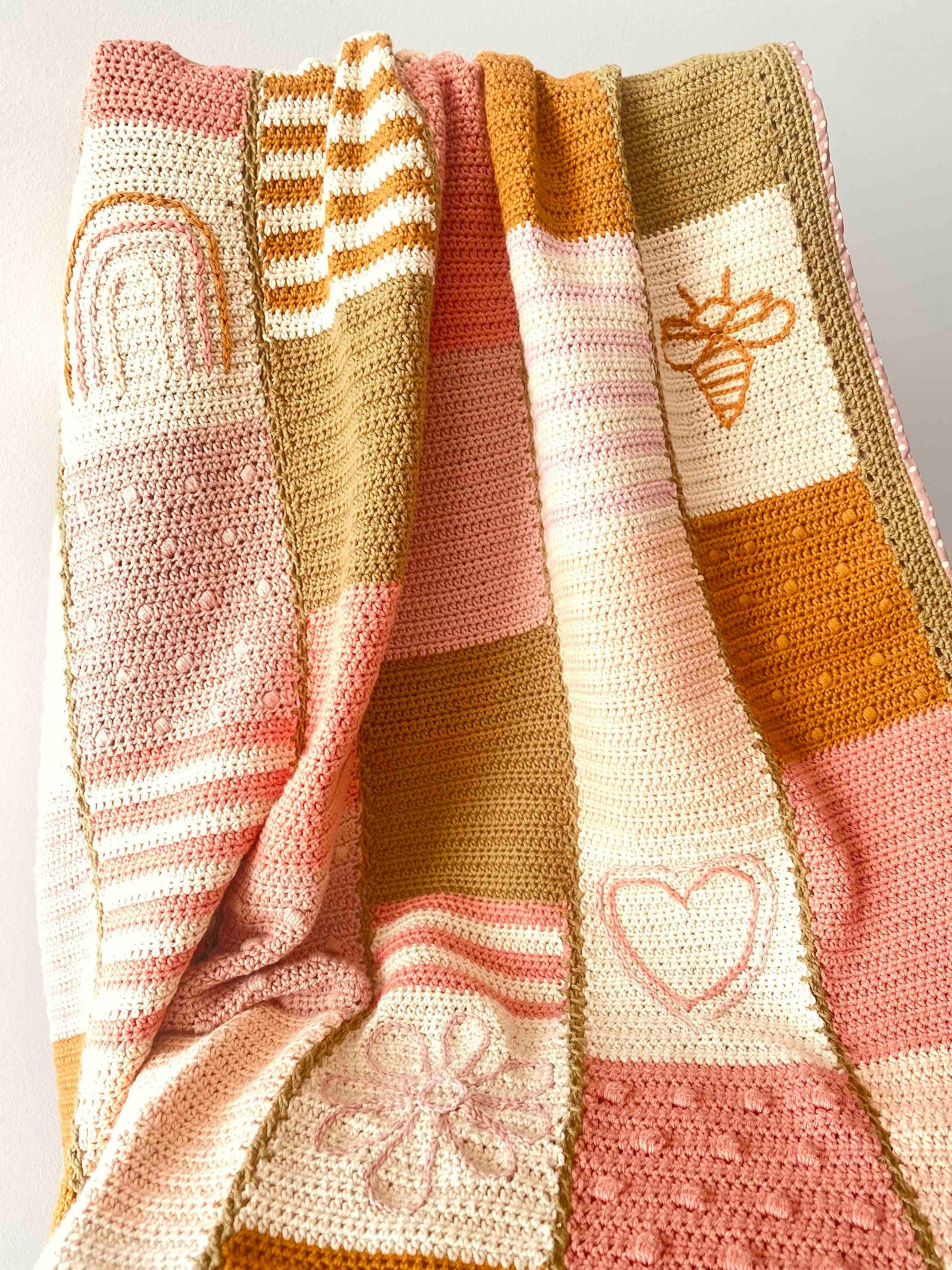 Crocheted baby blanket in a soft earth-tone patchwork design featuring hearts, bees, flowers, and rainbows