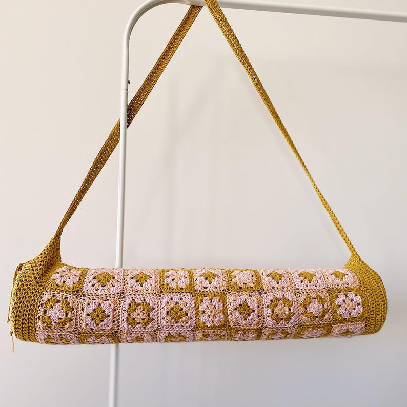 K&ouml;nnte beinhalten: Eine geh&auml;kelte Yogamatte-Tasche mit einem rosa und senfgelben Muster. Die Tasche hat einen langen Riemen zum Tragen &uuml;ber der Schulter.