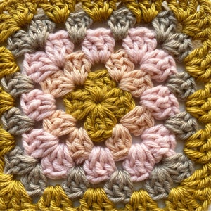 K&ouml;nnte beinhalten: Nahaufnahme eines geh&auml;kelten Quadrats mit einem zentralen Motiv aus rosa, pfirsichfarbenen und goldenen Blumen. Das Quadrat ist von einem Rand aus goldenen, braunen und grauen H&auml;kelmaschen umgeben.