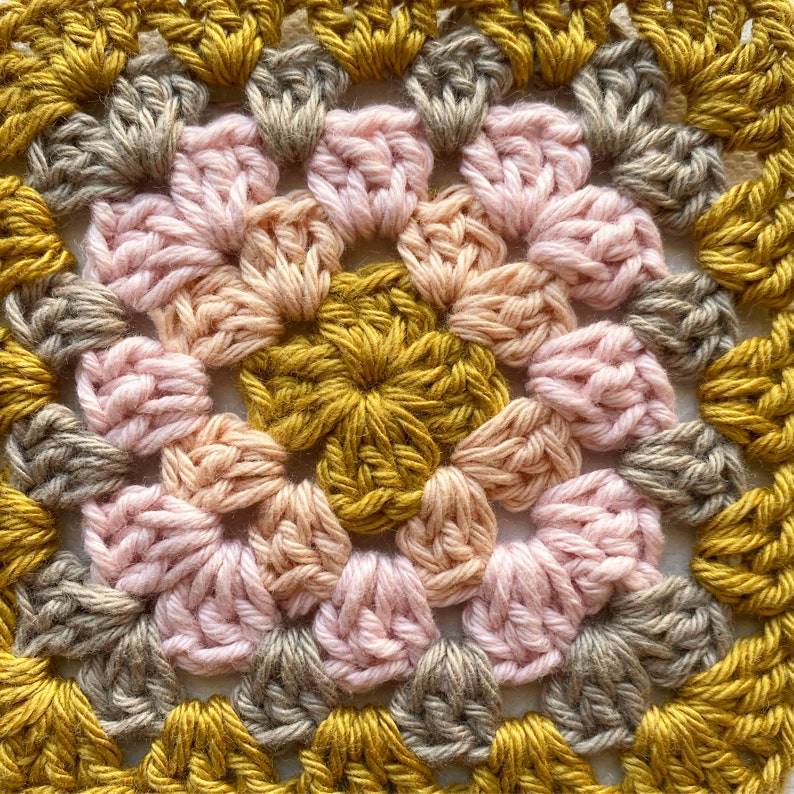 K&ouml;nnte beinhalten: Nahaufnahme eines geh&auml;kelten Quadrats mit einem zentralen Motiv aus rosa, pfirsichfarbenen und goldenen Blumen. Das Quadrat ist von einem Rand aus goldenen, braunen und grauen H&auml;kelmaschen umgeben.